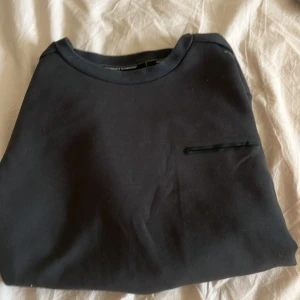 Svart sweatshirt med fickdetalj - En stilren svart sweatshirt med rund hals och en diskret fickdetalj på bröstet. Tröjan har långa ärmar och är gjord i mjukt bomullsmaterial, perfekt för en clean och enkel look. Passar dig som gillar minimalistisk stil.