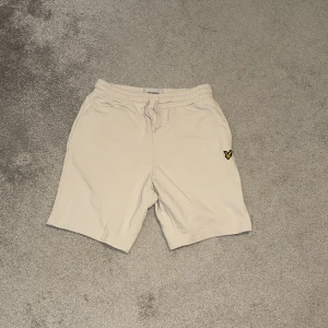 Beige shorts från Lyle & Scott - Beiga shorts från Lyle & Scott i mjuk bomull med elastisk midja och snörning. Klassisk passform med fickor på sidorna och en bakficka. Ikonisk gul logga på vänster ben. Perfekta för chill dagar eller när du vill vara bekväm.