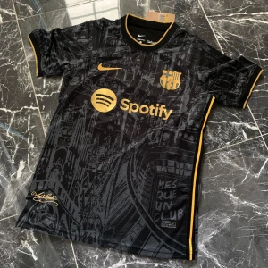 FC Barcelona svart matchtröja Nike - Snygg FC Barcelona fotbollströja i svart med guldiga detaljer och tryck från Nike. Tröjan har korta ärmar, gul Nike-logga, klubbmärke och Spotify som sponsor framtill. Unikt stadsmönster över hela tröjan och UNHCR-tryck på ryggen. Tillverkad i lätt och ventilerande material.