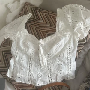 Vit broderad blus med knyt - Superfin vit blus med broderade blommor och spetsdetaljer. Korta puffärmar och söt knytning framtill. Perfekt till sommaren och ger en romantisk vibe till outfiten.
