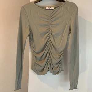Ribbad topp - NA-KD - Trendig olivgrön långärmad topp från NA-KD med ribbad struktur och snygg scrunch framtill. Modellen har en smickrande passform och vågig kant nertill💚ALDRIG ANVÄND 