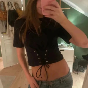 Svart  topp med snörning - Snygg svart  topp med korta ärmar och cool snörningsdetalj framtill. T-shirten har rund halsringning och snörningen ger en edgy vibe. Perfekt att matcha med jeans eller kjol för en trendig look.