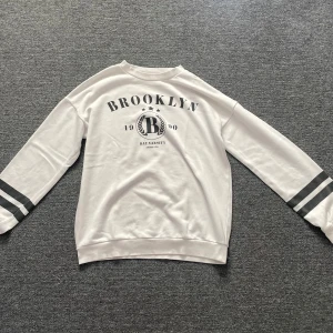 Vit sweatshirt Brooklyn H&M - Vit sweatshirt från H&M med svart Brooklyn-tryck framtill och två svarta ränder på ärmarna. Skön passform med rund hals och långa ärmar. Perfekt för en avslappnad streetstyle-look.