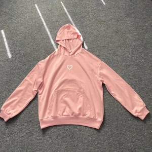 Rosa hoodie med hjärta från Shein - Söt ljusrosa hoodie från Shein med vit hjärt-detalj på bröstet. Tröjan har huva, magficka och långa ärmar. Perfekt för dig som gillar en chill och gullig stil. Mjuk och skön, passar till jeans eller leggings.