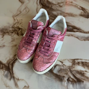 Valentino Garavani rosa glittriga sneakers - Snygga sneakers från Valentino Garavani i rosa glitter med vita skinnpaneler och rosa snörning. Skorna har rund tå, platt sula och coola nitar baktill. Perfekta för dig som vill sticka ut med extra bling och exklusiv känsla.