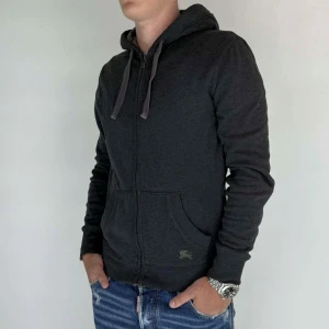Burberry Zip Hoodie  - Nu har vi nöjet att erbjuda en exklusiv zip hoodie från det välrenommerade varumärket Burberry. Hoodien är i storlek Medium och är i mycket gott skick, utan några synliga defekter. Modellen på bilden är 180 cm lång och väger 75 kg, vilket kan ge en god indikation på plaggets passform.