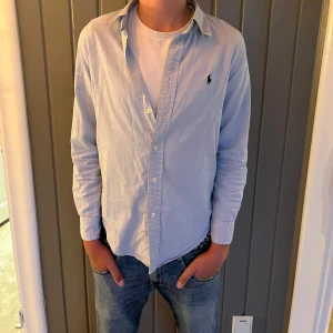 Ljusblå skjorta från Ralph Lauren - Klassisk ljusblå skjorta från Ralph Lauren med broderad logga på bröstet. Skjortan har lång ärm, knappar framtill och normal passform. Perfekt att bära öppen över en t-shirt eller stängd för en mer clean look.