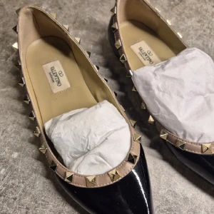 Valentino Rockstud ballerinaskor svart - Snygga Valentino Rockstud ballerinaskor i svart lackat skinn med spetsig tå. Skorna har beige kant med ikoniska guldiga nitar runtom för en edgy vibe. Perfekta för dig som vill sticka ut med lyxig designerstil och coola detaljer.