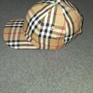 Burberry rutig keps i bomull - Snygg keps från Burberry i klassiskt beige rutigt mönster med svarta, vita och röda detaljer. Justerbart spänne baktill med guldfärgad detalj och broderad logga. Tillverkad i bomull och fodrad med svart tyg där Burberry-loggan syns.