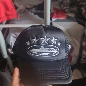 Corteiz keps - Snygg svart keps med mesh på sidorna och broderade stjärnor framtill och på sidan. Framsidan har även ett broderat motiv av en bil i silver. Justerbar snapback baktill för perfekt passform. Perfekt för dig som vill sticka ut med en cool accessoar.