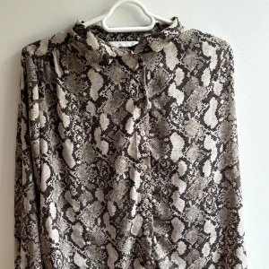 Ormmönstrad skjorta från H&M, strl 40 - Snygg skjorta från H&M i storlek 40 med ormmönster i beige och svart. Skjortan har klassisk krage, knäppning framtill och långa ärmar. Perfekt för dig som vill sticka ut med ett djurmönster och gillar trendiga detaljer.