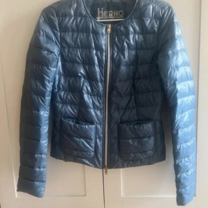 Blå dunjacka från Herno - Snygg blå dunjacka från Herno med quiltad design och rund halsringning. Jackan har dragkedja framtill, två fickor och diskret logga på ärmen. Perfekt för dig som vill ha en lätt och stilren jacka till vardags. Passar lika bra till jeans som till kjol.