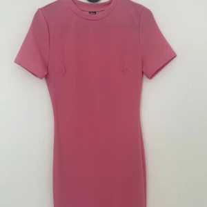 Rosa kortklänning från SHEIN XS - Enkel och snygg rosa kortklänning från SHEIN i storlek XS. Klänningen har korta ärmar, rund halsringning och en liten slits nedtill. Tillverkad i ett mjukt och stretchigt syntetmaterial som sitter slim på kroppen.