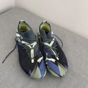 Puma fotbollsskor i blått,lime och vit - Snygga fotbollsskor från Puma med coolt grafiskt mönster i blått, svart, lime och vitt. Skorna har snörning, stickad strumpliknande krage och greppvänlig sula med dobbar. Perfekta för dig som vill sticka ut på planen!