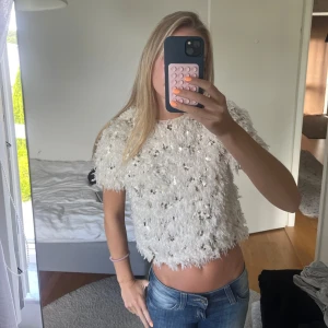 Vit glittrig topp med paljetter - Superfin vit topp med kort ärm och glittriga paljetter över hela. Toppen har en fluffig, nästan fjäderliknande textur och är croppad i modellen. Perfekt för dig som vill sticka ut och glänsa lite extra.