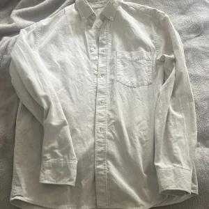 Vit manchesterskjorta med knappar - Vit skjorta i manchestertyg med klassisk krage och knappstängning hela vägen. Skjortan har en bröstficka och långa ärmar med manschett. Tunt randigt mönster i tyget ger en diskret struktur. Perfekt för dig som gillar stilrena och enkla plagg.