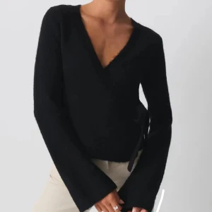 Svart omlottstickad tröja med v-ringning - Säljer en svart stickad tröja med omlottdesign och djup v-ringning. Tröjan har långa ärmar med lätt utsvängda ärmslut och knytning i sidan för en snygg detalj. Perfekt för lager på lager och en stilren look.