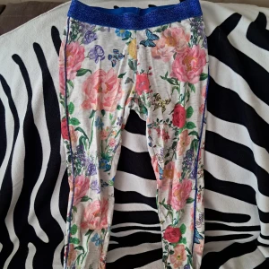 Blommiga leggings med blått glitterband - Superfina leggings med färgglatt blommönster och fjärilar på vit botten. Midjan har ett blått glitterresår som ger extra stilpoäng. Den är för barn- storlek 140, för ålder 9-10 år.