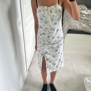 Vit blommig klänning med slits - Supersöt vit klänning med blått blommönster, smala axelband och knappar framtill. Klänningen har rynkade detaljer längs sidorna, en snygg slits framtill och en feminin passform. Perfekt för sommarens alla tillfällen!