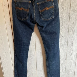 Mörkblå jeans från Nudie Jeans - Snygga mörkblå jeans från Nudie Jeans med orangea sömmar och vågdetaljer på bakfickorna. Jeansen har normal passform. Perfekta för dig som gillar en tidlös och clean look. Storlek W29 L34, endast testade.