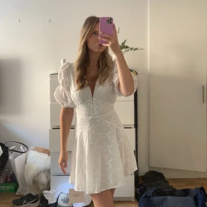 Vit kort klänning med puffärmar - Supersöt vit kort klänning med puffärmar och djup v-ringning. Klänningen har en markerad midja och diskret blommigt mönster i tyget. Perfekt för student! Kan möjligtvis ha någon liten fläck på sig!