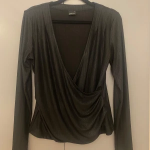 Svart omlottopp från Gina Tricot - Snygg svart omlottopp från Gina Tricot med djup v-ringning och långa ärmar. Toppen har ett subtilt glansigt tyg med diskreta vertikala ränder och draperad effekt framtill. Perfekt för dig som gillar stilrena och trendiga plagg.