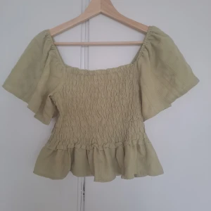 Beige blus med volang och puffärm - Supersöt beige blus med smockad midja, volang nertill och vida, korta puffärmar. Blusen har en fyrkantig ringning och är gjord i ett lätt, luftigt tyg som känns perfekt för varmare dagar.