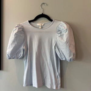 Ljusblå topp med puffärmar H&M - Supersöt ljusblå topp från H&M med puffiga korta ärmar och rund halsringning. Ärmarna har en ballongformad look och resår vid avslutet. Perfekt för dig som vill ha en enkel men trendig stil med extra volym på axlarna.