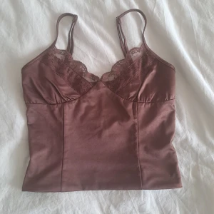 Brun satin linnetopp med spets - Supersnygg brun linnetopp i satin med tunna axelband och v-ringning. Toppen har fina spetsdetaljer längs bysten som ger en romantisk vibe. Perfekt att styla med jeans eller kjol för en trendig look.