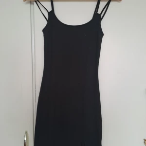 Svart kort klänning med slits - En svart, tight kortklänning med dubbla smala axelband och en liten slits framtill. Klänningen har en enkel och stilren design som sitter figurnära och är perfekt för dig som gillar minimalistisk stil.