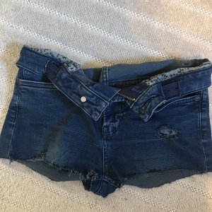 Mörkblå jeansshorts med en leopard detalj - Snygga mörkblå jeansshorts med mid waist och råa kanter. Shortsen har slitningar och en cool leopardmönstrad detalj vid ena benet. Klassisk femficksmodell med dragkedja och knapp. Perfekta för sommarens alla äventyr.