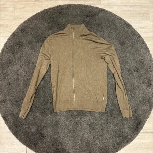 Beige kofta med dragkedja från Lindbergh - Snygg beige kofta från Lindbergh med hel dragkedja framtill och hög krage. Tröjan är stickad i ett tunt material och har långa ärmar samt ribbade muddar vid ärmslut och nederkant. Perfekt för lager-på-lager och stilrena outfits.