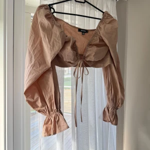 Beige croppad blus från Missguided - Supersnygg croppad blus från Missguided i beige bomull. Toppen har långa puffärmar med resår vid ärmslut, djup v-ringning och knyt framtill. Perfekt för dig som vill ha en trendig och luftig look med voluminösa ärmar och coola detaljer.