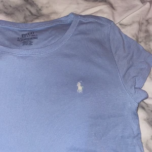 Ljusblå t-shirt från Polo Ralph Lauren - Ljusblå t-shirt från Polo Ralph Lauren med klassisk vit broderad logga på bröstet. Använd en gång! Passar en Xs och S