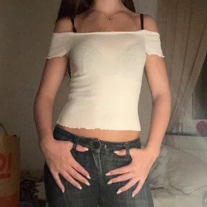 Vit ribbad croppad topp - Säljer en vit ribbad topp med croppad passform. Toppen har stretchigt material och kan bäras både offshoulder och med vanliga axelband. Den har vågig kant nertill och vid ärmslut, vilket ger en söt och trendig look. Perfekt till jeans eller kjol!