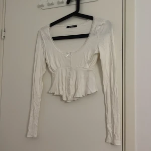 Vit volangblus från Gina Tricot XS - Supersöt vit blus från Gina Tricot med lång ärm, volang nertill och spetsdetaljer längs urringningen. Söt liten rosett framtill och croppad passform. Perfekt för dig som gillar romantisk och feminin stil.
