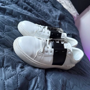 Valentino Garavani Open sneakers vit/svart - Snygga Valentino Garavani Open sneakers i vitt skinn med bred svart läderdetalj över snörningen. Klassisk rund tå, platt sula och ikoniska nitar baktill. Perfekta för dig som gillar stilrena och lyxiga sneakers med en twist.
