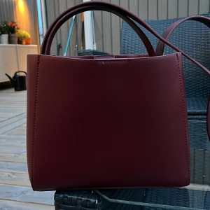 Vinröd handväska från Gina Tricot - Snygg vinröd handväska från Gina Tricot i stilren boxig modell. Väskan har både handtag och axelrem, samt en innerficka med dragkedja. Tillverkad i slätt syntetmaterial och har coola silverfärgade detaljer på sidorna. Säljer denna då den inte kommer till användning. Den är köpt i hösten 2024 och använd enstaka gånger 