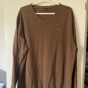 Brun stickad tröja Hampton Republic XL - Snygg brun stickad tröja från Hampton Republic i storlek XL. Tröjan har v-ringad hals och en liten broderad logga på bröstet. Perfekt för lager-på-lager och passar till både jeans och chinos. Mjuk och skön känsla med långa ärmar och ribbade muddar.
