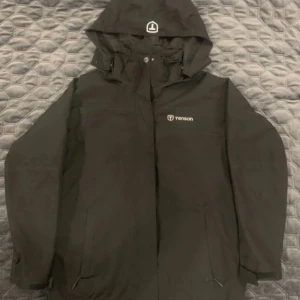 Svart vindjacka från Tenson med huva - Svart vindjacka från Tenson med justerbar huva och dragkedja framtill. Jackan har diskret logga på bröstet och meshfoder på insidan för extra komfort. Perfekt för blåsiga dagar och enkel att matcha med allt.