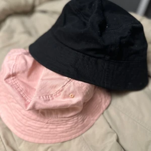 Svart och rosa bucket hat duo - Två stilrena bucket hats i svart och ljusrosa från lager 157. Klassisk form med mjuk brätte och sydda detaljer. Perfekta för att lyfta din look under soliga dagar. Tillverkade i bomull för en bekväm känsla. 20kr styck