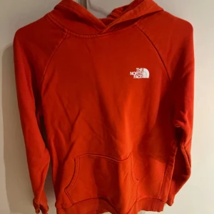 Röd hoodie från The North Face - Snygg röd hoodie från The North Face med vit logga på bröstet och stor logga med texten 'Never Stop Exploring' på ryggen. Tröjan har huva, magficka och ribbade muddar. Perfekt för dig som gillar streetwear och vill sticka ut. 