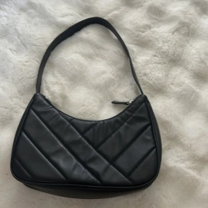 Svart quiltad axelväska från H&M - Snygg svart axelväska från H&M i quiltad design med diagonala sömmar. Väskan har en mjuk, rundad form och är gjord i syntetmaterial. Stängs med dragkedja upptill och har ett stilrent, minimalistiskt uttryck som passar till många outfits.