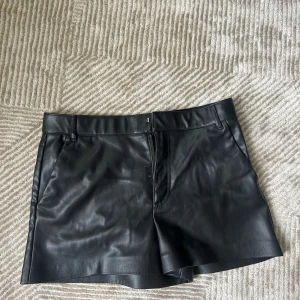 Svarta skinnshorts med dragkedja - Snygga svarta shorts i skinnimitation med klassisk passform och dragkedja framtill. De har bälteshällor och en stilren look som passar perfekt till en trendig outfit. Perfekta för dig som vill sticka ut med en edgy vibe.