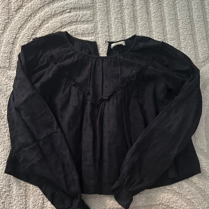 Mörkblå blus med volangdetalj - Mörkblå blus i bomull med diskret prickigt mönster och volangdetalj framtill. Långärmad modell med lätt rynkade axlar och rund halsringning. Blusen har en liten öppning bak i nacken för en extra touch.