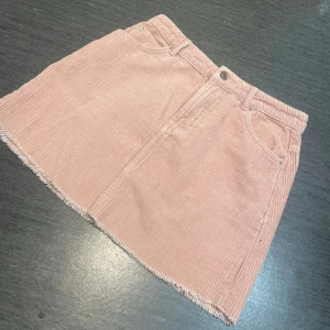 Rosa manchesterkjol från Gina Tricot - Supersöt rosa kortkjol i manchester från Gina Tricot. Kjolen har klassiska fickor fram, dragkedja och knapp, samt rå kant nertill för en chill vibe. Perfekt för dig som vill ha en trendig och bekväm kjol med lite retrofeeling.