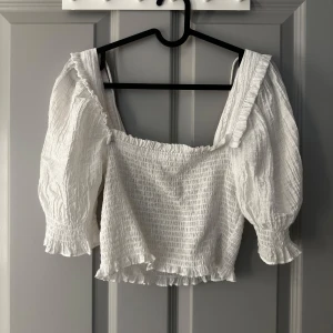 Vit topp med puffärm och volang - Supersöt vit topp med smockad struktur, puffiga trekvartsärmar och volangdetaljer vid ärmslut och nederkant. Fyrkantig ringning med volangkant och knytband i nacken. 