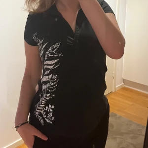 Svart T-shirt med vit växtprint Lindex - Svart T-shirt från Lindex med vit växtinspirerad print på ena sidan. Modellen har korta ärmar, krage och lätt rynk vid bysten för en snygg detalj. Skön och mjuk bomullskänsla, perfekt till jeans eller kjol. Axelbredd: 37cm. Längd: 57cm. Byst: 47cm.