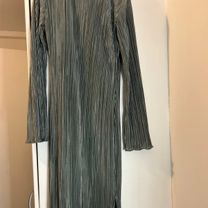 Plisserad silverklänning från SHEIN - Snygg lång plisserad klänning från SHEIN i metallic silver. Klänningen har långa ärmar, rak passform och en liten slits nertill. Materialet är glansigt och ger en cool, modern vibe. Perfekt för dig som vill sticka ut med stil.