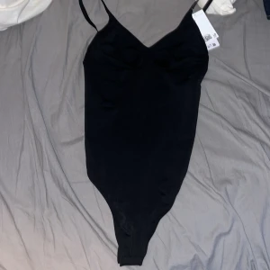 Svart bodysuit med smala axelband - Svart bodysuit med smala justerbara axelband och v-ringad urringning. Snygg rynkdetalj vid bysten och figurnära passform. Perfekt att styla med jeans eller kjol. Tyvärr satt den för tight runt brösten. Dm för lägre pris💗
